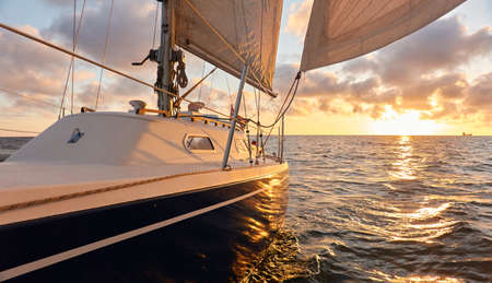 Yacht sailing in an open sea at sunset. Top down view of the deck, mast and sails. Clear sky after the rain, dramatic glowing clouds, golden sunlight. Waves, water splashes. Travel, cruise, sportの写真素材