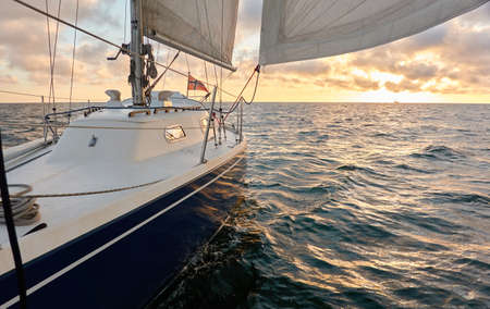 Yacht sailing in an open sea at sunset. Top down view of the deck, mast and sails. Clear sky after the rain, dramatic glowing clouds, golden sunlight. Waves, water splashes. Travel, cruise, sportの写真素材