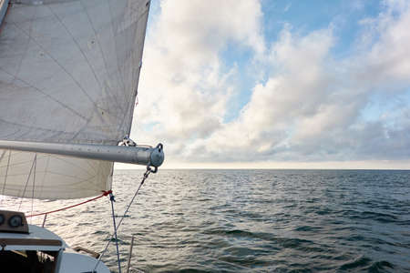 White sloop rigged yacht sailing in an open sea at sunset. Clear sky after the rain, dramatic glowing clouds, golden sunlight. Waves, water splashes. Travel, cruise, sport, adventure, wanderlustの写真素材