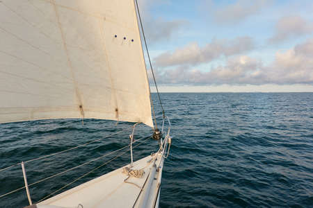 White sloop rigged yacht sailing in an open sea at sunset. Clear sky after the rain, dramatic glowing clouds, golden sunlight. Waves, water splashes. Travel, cruise, sport, adventure, wanderlustの写真素材
