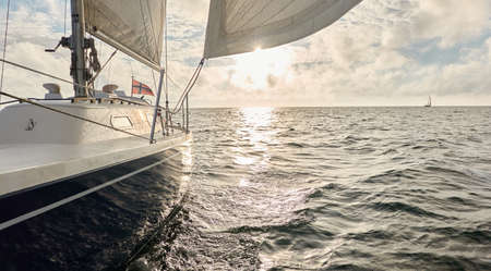 Yacht sailing in an open sea at sunset. Top down view of the deck, mast and sails. Clear sky after the rain, dramatic glowing clouds, golden sunlight. Waves, water splashes. Travel, cruise, sportの写真素材