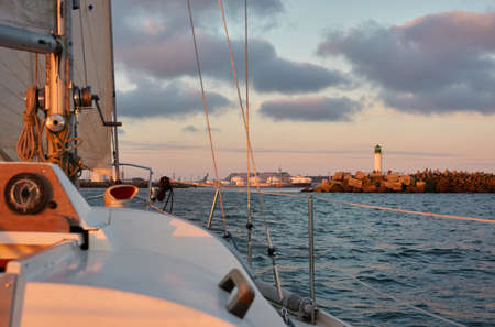 Yacht sailing in the Baltic sea at sunset. View from the cockpit. Lighthouse. Clear sky after the rain, dramatic glowing clouds, golden sunlight. Waves, water splashes. Travel, cruise, sportの写真素材