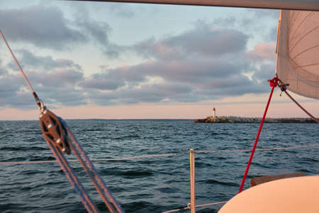 White sloop rigged yacht sailing in an open sea at sunset. Clear sky after the rain, dramatic glowing clouds, golden sunlight. Waves, water splashes. Travel, cruise, sport, adventure, wanderlustの写真素材