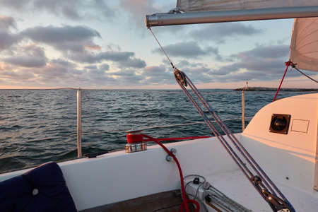 Yacht sailing in the Baltic sea at sunset. View from the cockpit. Lighthouse. Clear sky after the rain, dramatic glowing clouds, golden sunlight. Waves, water splashes. Travel, cruise, sportの写真素材