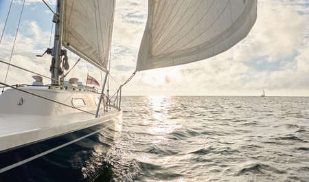 Yacht sailing in an open sea at sunset. Top down view of the deck, mast and sails. Clear sky after the rain, dramatic glowing clouds, golden sunlight. Waves, water splashes. Travel, cruise, sportの写真素材
