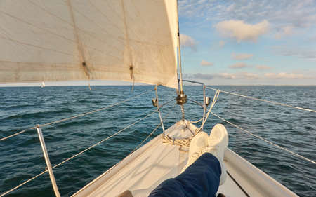 White sloop rigged yacht sailing in an open sea at sunset. Clear sky after the rain, dramatic glowing clouds, golden sunlight. Waves, water splashes. Travel, cruise, sport, adventure, wanderlustの写真素材