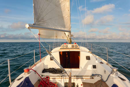 White sloop rigged yacht sailing in an open sea at sunset. Clear sky after the rain, dramatic glowing clouds, golden sunlight. Waves, water splashes. Travel, cruise, sport, adventure, wanderlustの写真素材