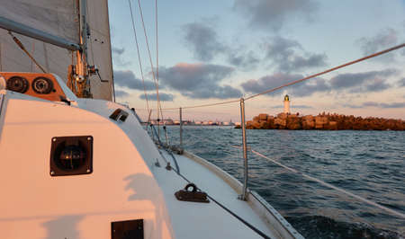 Yacht sailing in the Baltic sea at sunset. View from the cockpit. Lighthouse. Clear sky after the rain, dramatic glowing clouds, golden sunlight. Waves, water splashes. Travel, cruise, sportの写真素材