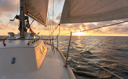 Yacht sailing in an open sea at sunset. Top down view of the deck, mast and sails. Clear sky after the rain, dramatic glowing clouds, golden sunlight. Waves, water splashes. Travel, cruise, sportの写真素材