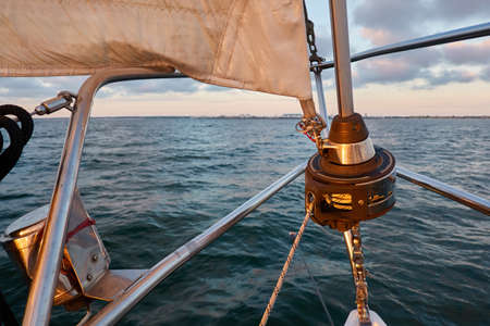 White sloop rigged yacht sailing in an open sea at sunset. Clear sky after the rain, dramatic glowing clouds, golden sunlight. Waves, water splashes. Travel, cruise, sport, adventure, wanderlustの写真素材