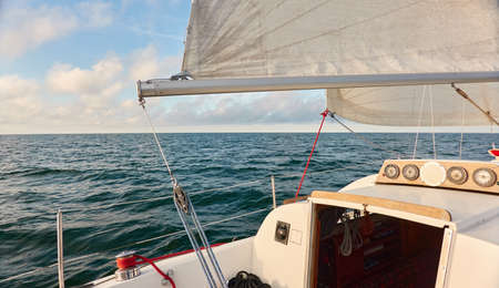 White sloop rigged yacht sailing in an open sea at sunset. Clear sky after the rain, dramatic glowing clouds, golden sunlight. Waves, water splashes. Travel, cruise, sport, adventure, wanderlustの写真素材