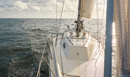 White sloop rigged yacht sailing in an open sea at sunset. Clear sky after the rain, dramatic glowing clouds, golden sunlight. Waves, water splashes. Travel, cruise, sport, adventure, wanderlustの写真素材