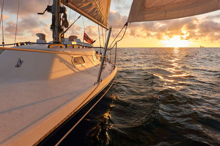 Yacht sailing in an open sea at sunset. Top down view of the deck, mast and sails. Clear sky after the rain, dramatic glowing clouds, golden sunlight. Waves, water splashes. Travel, cruise, sportの写真素材