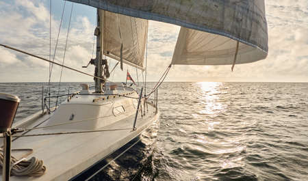 Yacht sailing in an open sea at sunset. Top down view of the deck, mast and sails. Clear sky after the rain, dramatic glowing clouds, golden sunlight. Waves, water splashes. Travel, cruise, sportの写真素材