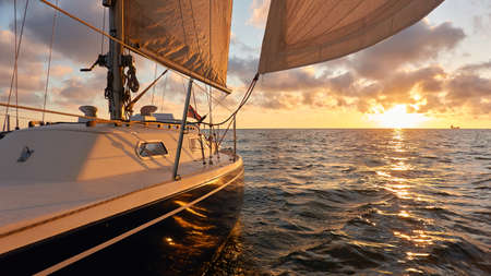 Yacht sailing in an open sea at sunset. Top down view of the deck, mast and sails. Clear sky after the rain, dramatic glowing clouds, golden sunlight. Waves, water splashes. Travel, cruise, sportの写真素材
