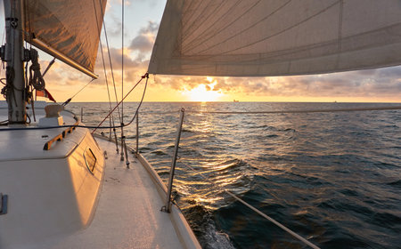 Yacht sailing in an open sea at sunset. Top down view of the deck, mast and sails. Clear sky after the rain, dramatic glowing clouds, golden sunlight. Waves, water splashes. Travel, cruise, sportの写真素材