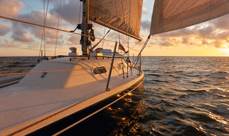 Yacht sailing in an open sea at sunset. Top down view of the deck, mast and sails. Clear sky after the rain, dramatic glowing clouds, golden sunlight. Waves, water splashes. Travel, cruise, sportの写真素材