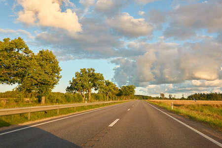 Highway (new asphalt road) through the agricultural field and forest at sunset. Dramatic sky after the thunderstorm. Summer, early autumn. Vacations, adventure, road trip, remote places. View from carの写真素材
