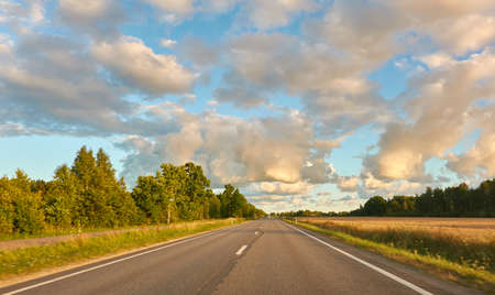 Highway (new asphalt road) through the agricultural field and forest at sunset. Dramatic sky after the thunderstorm. Summer, early autumn. Vacations, adventure, road trip, remote places. View from carの写真素材