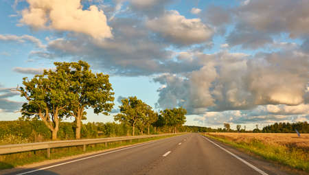 Highway (new asphalt road) through the agricultural field and forest at sunset. Dramatic sky after the thunderstorm. Summer, early autumn. Vacations, adventure, road trip, remote places. View from carの写真素材