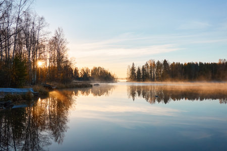 Panoramic view of the forest lake at sunrise. Soft golden sunlight, clear blue sky, reflections on water, fog, frost. Mighty trees. Finland. Nature, seasons, ecology, environment, eco tourism themesの写真素材