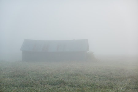 Lonely wooden farm house or shed (log cabin) in a thick fog at sunrise. Forest lawn, meadow, overgrown agricultural field. Atmospheric autumn landscape. Finlandの写真素材