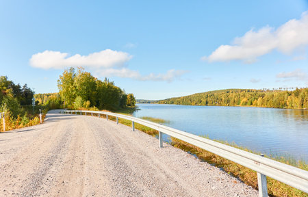 Gravel road (highway) near the forest lake. Sunny day. Concept autumn landscape. Transportation, logistics, freedom, travel, tourism, wanderlust, the way forward themesの写真素材