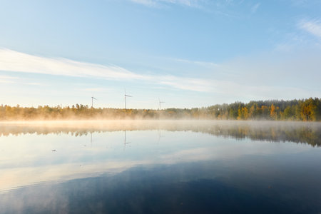 Forest lake in a morning fog. Wind farm, wind turbine generators. Clear blue sky, sunshine. Finland. Scandinavia, Europe. Autumn landscape. Nature, ecology, alternative and renewable energyの写真素材