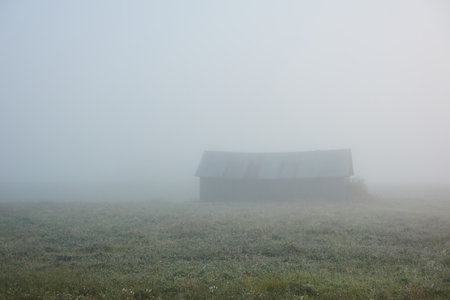 Lonely wooden farm house or shed (log cabin) in a thick fog at sunrise. Forest lawn, meadow, overgrown agricultural field. Atmospheric autumn landscape. Finlandの写真素材