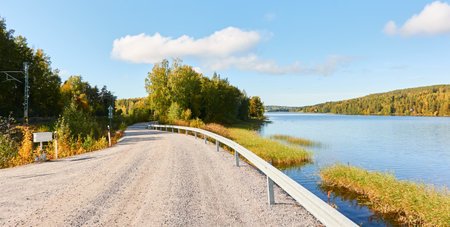 Gravel road (highway) near the forest lake. Sunny day. Concept autumn landscape. Transportation, logistics, freedom, travel, tourism, wanderlust, the way forward themesの写真素材