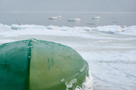 Green rowing fishing boat standing on land. Frozen Baltic seashore. Snow, ice, cloudy day. Concept winter landscape. Nature, environment, ecology, pollution themesの写真素材