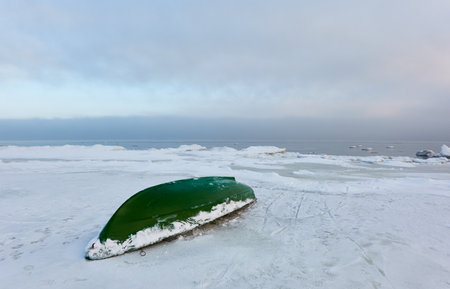 Green rowing fishing boat standing on land. Frozen Baltic seashore. Snow, ice, cloudy day. Concept winter landscape. Nature, environment, ecology, pollution themesの写真素材