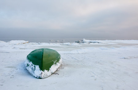 Green rowing fishing boat standing on land. Frozen Baltic seashore. Snow, ice, cloudy day. Concept winter landscape. Nature, environment, ecology, pollution themesの写真素材