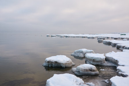 A view from the frozen snow-covered Baltic sea shore. cloudy day. Crystal clear water, cracked ice. Fickle weather, snowstorm, blizzard. Winter tourism, global warming, climate change, ecology themesの写真素材
