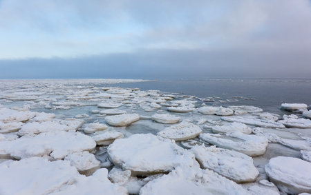 A view from the frozen snow-covered Baltic sea shore. cloudy day. Crystal clear water, cracked ice. Fickle weather, snowstorm, blizzard. Winter tourism, global warming, climate change, ecology themesの写真素材