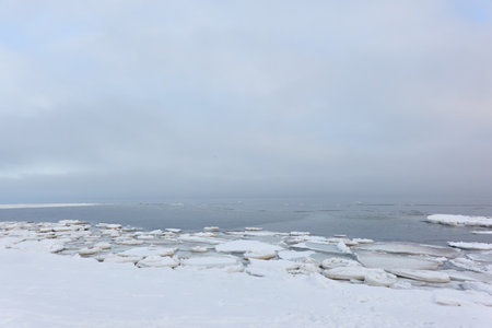 A view from the frozen snow-covered Baltic sea shore. cloudy day. Crystal clear water, cracked ice. Fickle weather, snowstorm, blizzard. Winter tourism, global warming, climate change, ecology themesの写真素材