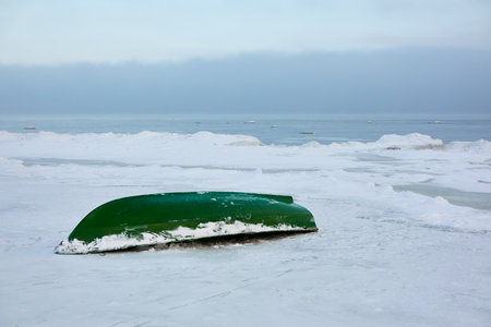 Green rowing fishing boat standing on land. Frozen Baltic seashore. Snow, ice, cloudy day. Concept winter landscape. Nature, environment, ecology, pollution themesの写真素材