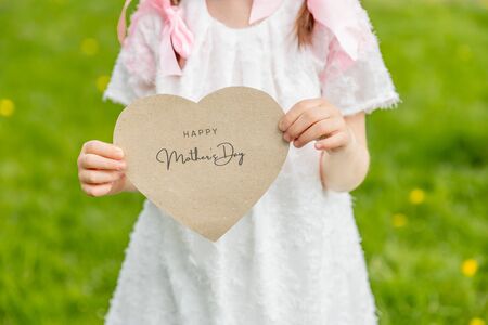 Happy Mother's Day written on a heart-shaped card held by a girl on grass backgroundの写真素材