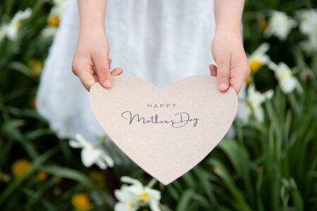 Happy Mother's Day written on a heart-shaped card held by a girl on flower and grass backgroundの写真素材