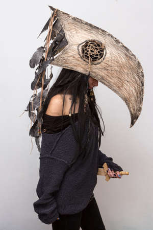 Beautiful girl dressed in witch or shaman halloween costume with black feathers and crow head on the white background with shadowsの写真素材