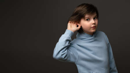 Curious young schoolboy 10-12 years old, holding hand at his ear, trying to overhear private conversation, dressed in casual clothes, posing at gray studio backgroundの写真素材