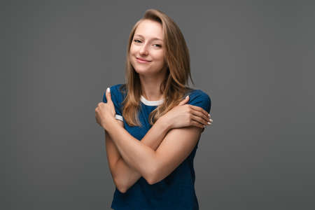Charming european blonde girl hugging herself hold shoulders, tilting head, smiling flirty romantic attitude, standing delighted gray background. Love, care tenderness conceptの写真素材