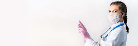 A young female doctor in a white coat, in a medical mask and pink sterile gloves hold syringe. Vaccination concept.の写真素材