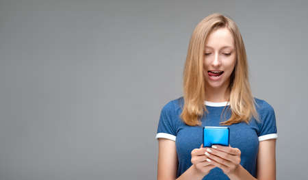 Horizontal shot of pleased Young woman with blond hair and a blue t shirt, uses mobile phone for...