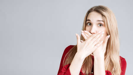Shocked young woman with long blonde hair dressed in casual clothes covering mouth with hands on gray background. Keep silence concept. Copy space for your textの写真素材