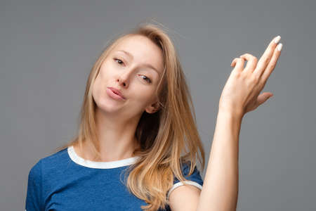 Funny young blonde female shoots in temple, tilts head, dressed in casual blue t shirt, demonstrates...