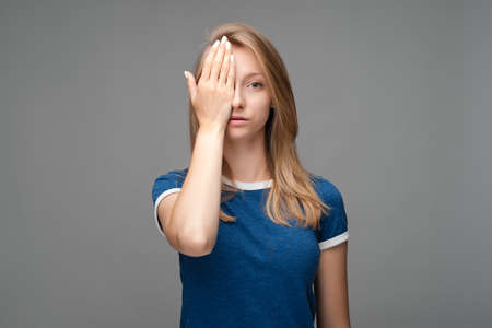 Photo of tired Young female with blond hair, covers face, feels fatigue, needs good rest, dressed in blue t shirt, isolated over gray background. Human emotions, facial expression concept.の写真素材