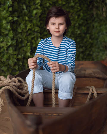 Young boy 10-12 years old in the striped shirt sits in a wooden boat. Studio shot. Concept of vacation and holidaysの写真素材