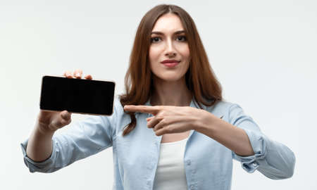 Young woman with long chestnut hair, dressed in casual clothes, Showing Smartphone Blank Screen,...