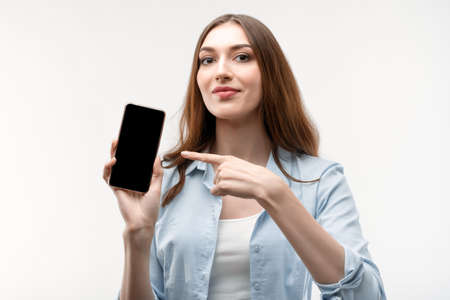 Young woman with long chestnut hair, dressed in casual clothes, Showing Smartphone Blank Screen,...